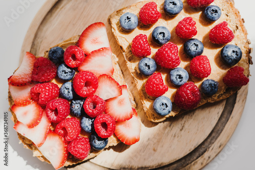 Two whole grain bread toast with peanut butter, fresh blueberry, raspberry and strawberry on wooden cutting board, top view. Breakfast toast with healthy topping