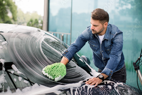 Handsome young Caucasian man cleaning his car windshield with green sponge mitten and soap foam outdoors at car wash service photo