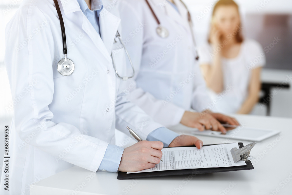Unknown woman-doctors at work with patient at the background. Female physicians filling up medical documents or prescription while standing in sunny hospital reception desk, close-up. Health care Unknown woman-doctors at work with patient at the background. Female physicians filling up medical documents or prescription while standing in sunny hospital reception desk, close-up. Health care