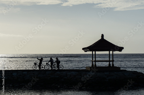 Silhouette of cyclists taking selfie near the sea. Cycling together with friends at the beach with flat sky at stone deck and balinese building