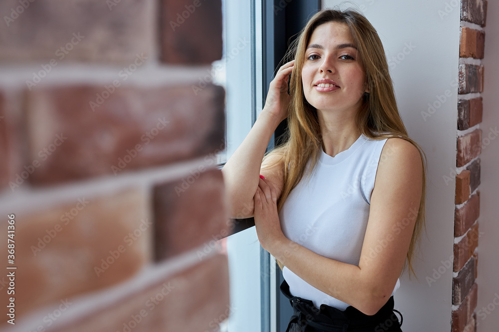portrait of young attractive caucasian woman posing at camera next to window, she looks at camera and smile. beautiful female with long hair stands near brickwall