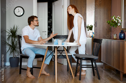 redhead woman is outraged that her husband works a lot, caucasian married couple quarrelling in the kitchen. man sit with laptop