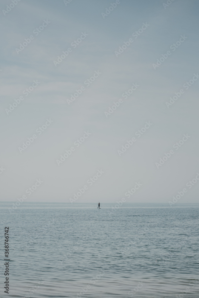 Minimal seascape with calm water and someone near the horizon line enjoying  the morning on a stand up paddle. Relaxing and fun outdoor summer activities for tourists. Seaside sport for active people.
