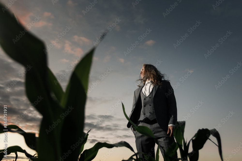 Obraz premium young stylish guy stands in a field in a gray suit at sunset. Blond with long hair in a cornfield. fashion concept