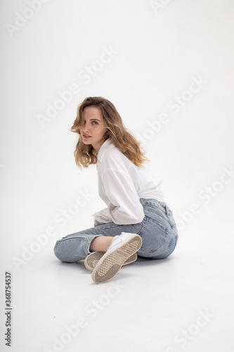 portrait of young pensive caucasian woman posing in shirt and blue jeans, sitting on white studio floor. model tests of pretty girl in basic clothes on cyclorama. attractive female poses, model tests