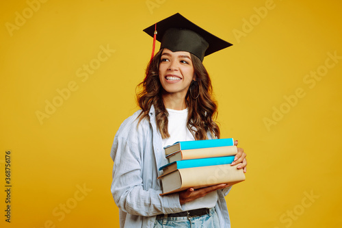 Bilde på lerret Graduate girl in a graduation hat on her head, with books stands on a yellow background