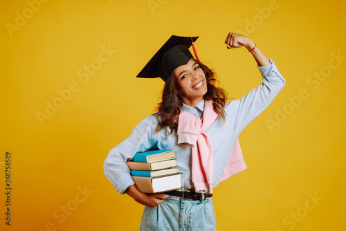 Billede på lærred Graduate girl in a graduation hat on her head, with books stands on a yellow background