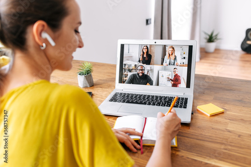 Back view of laptop display with a group of multiracial people on it, young woman watching online webinar and writing notes. Video call, online conference photo