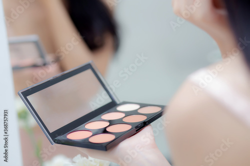 Beauty young asian woman happy and smile with face looking mirror applying makeup with brusher of cheek in the room, beautiful of girl holding brush make-up on facial, skin care and cosmetic concept.