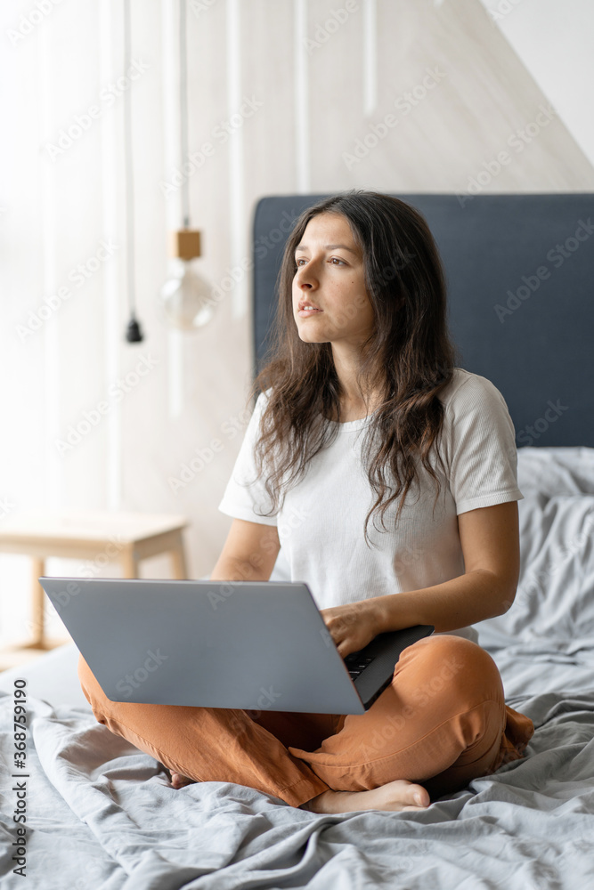 Beautiful young brunette girl with a laptop sitting on the bed. Stylish modern interior. A cozy workplace. Shopping on the Internet Beautiful young brunette girl with a laptop sitting on the bed. Stylish modern interior. A cozy workplace. Shopping on the Internet