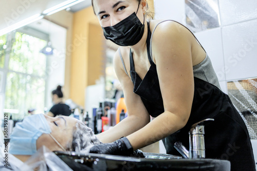 Hairdressing services during the coronavirus. Hands of hairdresser washing head of woman in mask. Hairdresser washing head of woman
