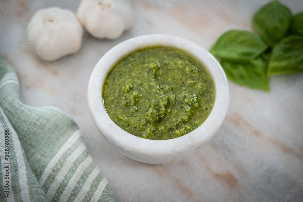 Photograph of fresh basil pesto, on the counter in a stoneware bowl, with garlic and basil leaves in the background with shallow depth of field