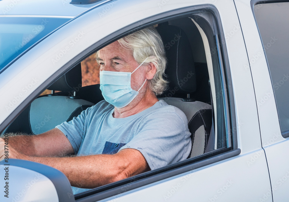 Attractive senior man driving his silver car wearing medical mask due to coronavirus happy for end lockdown at home Attractive senior man driving his silver car wearing medical mask due to coronavirus happy for end lockdown at home