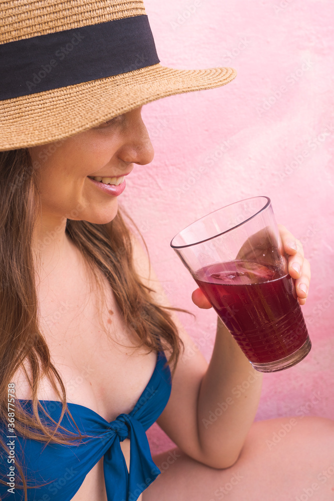 Beautiful blonde young woman smiling and holding a drink wearing a bikini in pink background Beautiful blonde young woman smiling and holding a drink wearing a bikini in pink background