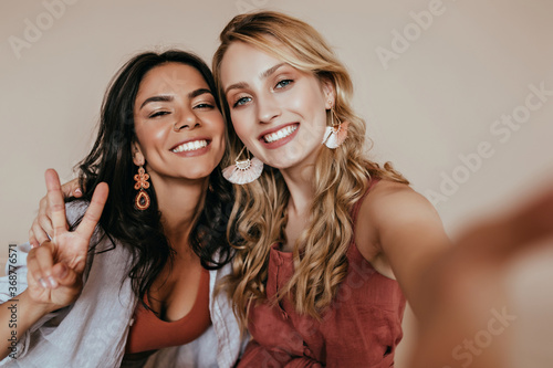 Pretty latin girl with dark hair posing with peace sign. Indoor photo of enthusiastic white woman making selfie with friend.
