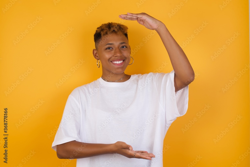 Young African American girl with afro short hair wearing white tshirt standing gesturing with hands showing big and large size sign, measure symbol. Smiling looking at the camera. Measuring concept. Young African American girl with afro short hair wearing white tshirt standing gesturing with hands showing big and large size sign, measure symbol. Smiling looking at the camera. Measuring concept.