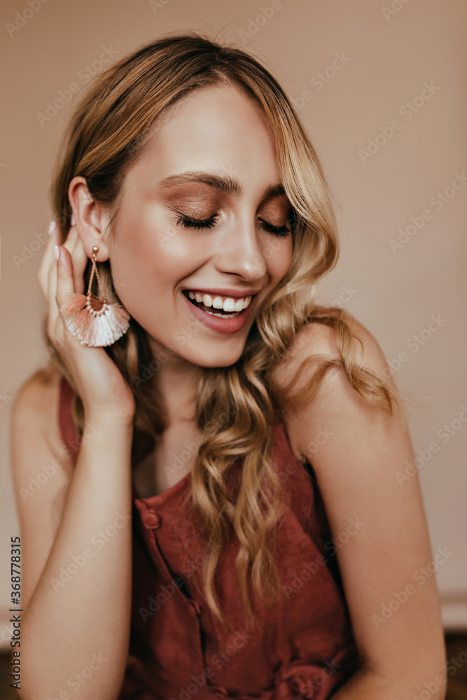 Pleasant caucasian woman with long wavy hair laughing with eyes closed. Fascinating stylish lady in earrings standing on light background. Pleasant caucasian woman with long wavy hair laughing with eyes closed. Fascinating stylish lady in earrings standing on light background.