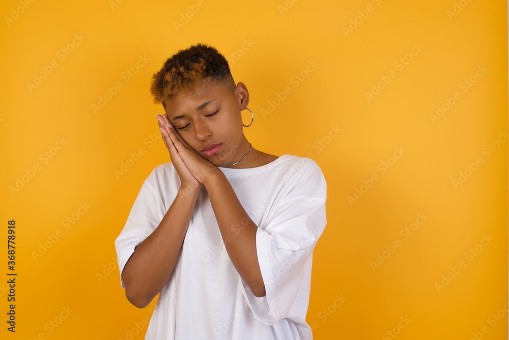 Young African American girl with afro short hair wearing white tshirt standing over isolated yellow wall sleeping tired dreaming and posing with hands together while smiling with closed eyes. Young African American girl with afro short hair wearing white tshirt standing over isolated yellow wall sleeping tired dreaming and posing with hands together while smiling with closed eyes.