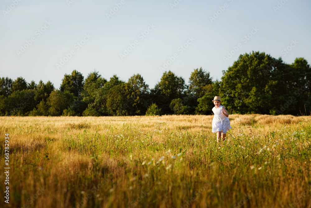 A happy and cheerful girl in a white sundress and hat runs across the field at sunset. A little girl runs in the summer on a rural field.