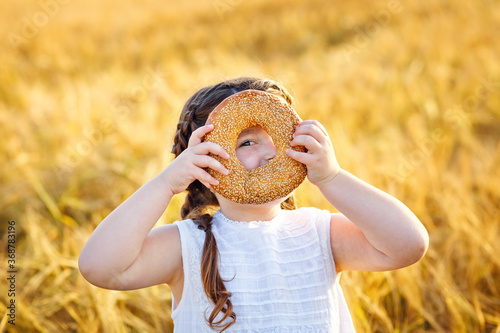 Fototapeta A smiling little girl in a white sundress and two pigtails on a wheat field with a bagel in the open air