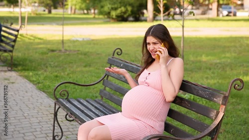 angry pregnant woman sitting on a bench in a summer city park and talking and cursing on her mobile phone