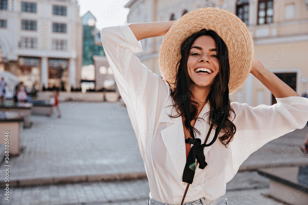 Charming european girl in white blouse expressing happiness in summer day. Outdoor photo of enthusiastic lady in glamorous hat standing on city background.