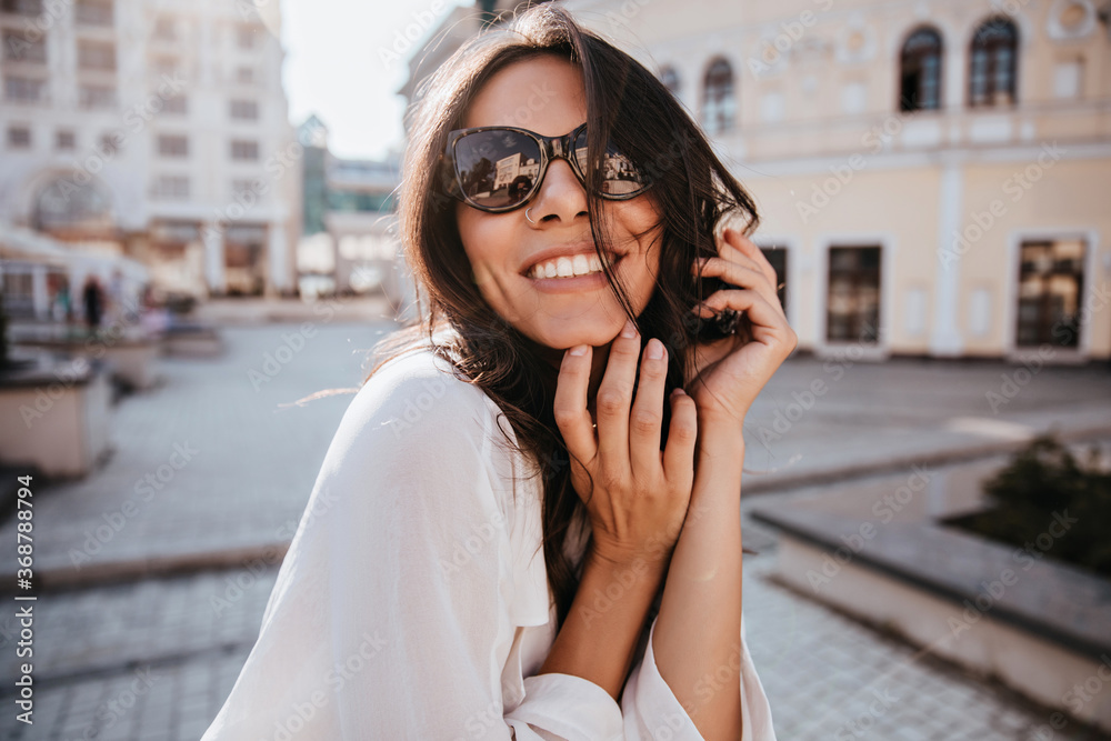 Pleased young woman in sunglasses posing with sincere smile on city background. Debonair european girl chilling in weekend.