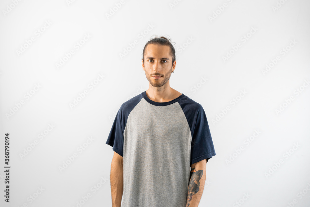 Hombre con pelo recogido, camiseta gris de mangas azules y tatuajes posa con rostro tranquilo en fondo blanco