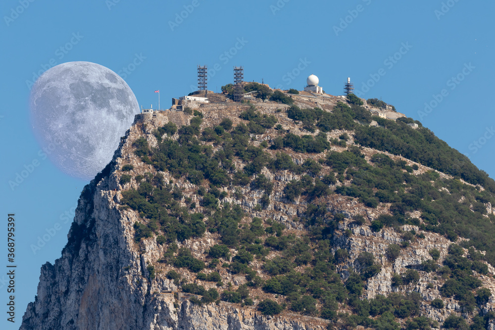 Telephoto of the northern summit of Gibraltar with a radio station in the sunshine and blue sky. The moon can be seen big behind the top of the huge rock.