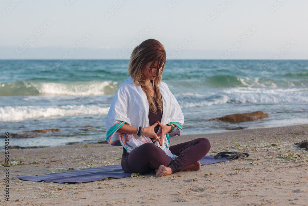 Young woman meditating while sitting on a yoga mat at the beach. Natural morning light shot with the Mediterranean Sea in the background.