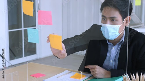 Normal view, medium shot, front view of young asian man in suit with facial mask thinking and writing on the post-it and put the sticky note to the transperant partion, in the office during DOVID-19 photo