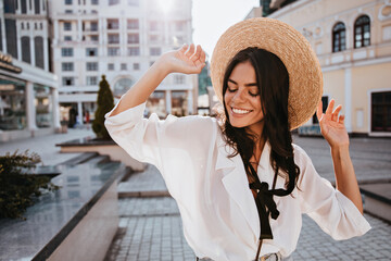 Good-humoured brunette lady posing on city background in hat. Outdoor portrait of attractive dark-haired girl standing on the street.