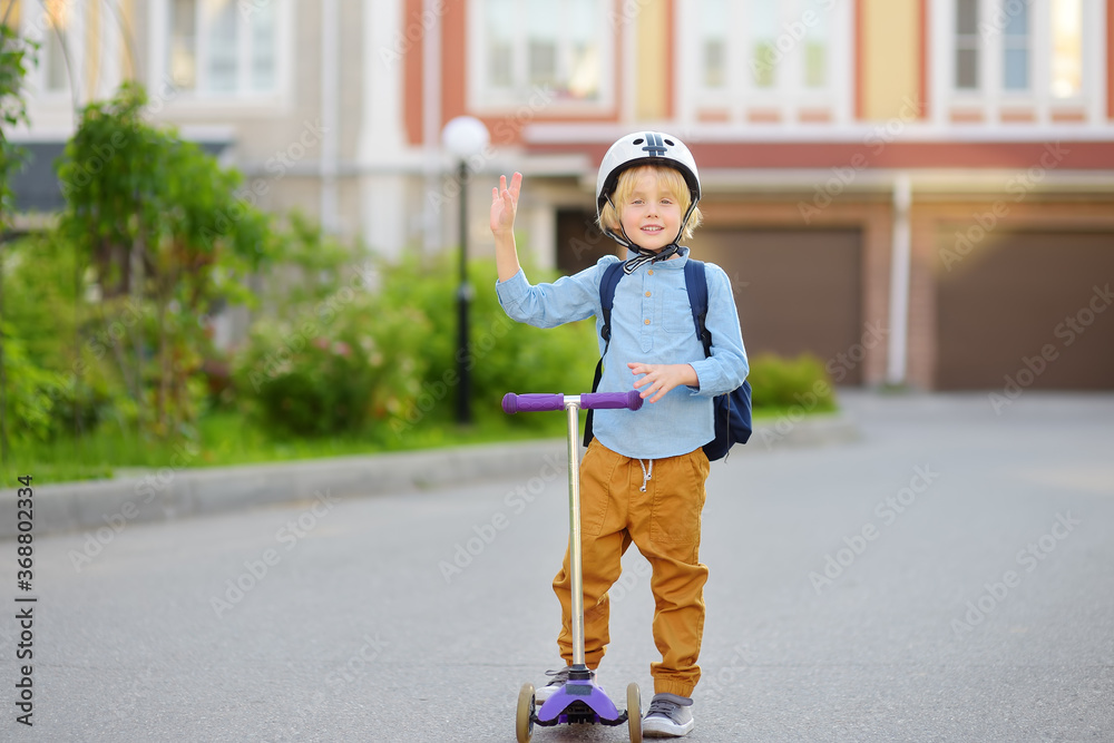 Little child in safety helmet riding scooter to school. Cute boy waving hand saying hi. Safety kids by way to school.