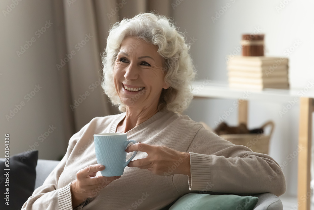 Untroubled 70s woman lean on couch drinking hot tea laughs feels carefree enjoy new day at calm retired life. No stress lifestyle, owner of modern cozy home, medical insurance healthy granny concept Untroubled 70s woman lean on couch drinking hot tea laughs feels carefree enjoy new day at calm retired life. No stress lifestyle, owner of modern cozy home, medical insurance healthy granny concept