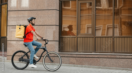 Fototapeta Naklejka Na Ścianę i Meble -  Delivery, work and sport. Young guy with beard in helmet with backpack rides a bicycle down the street
