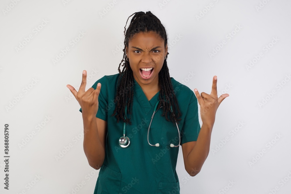 Born to rock this world. Joyful doctor woman in medical uniform screaming out loud and showing with raised arms horns or rock gesture, expressing excitement of being on concert of band. Born to rock this world. Joyful doctor woman in medical uniform screaming out loud and showing with raised arms horns or rock gesture, expressing excitement of being on concert of band.