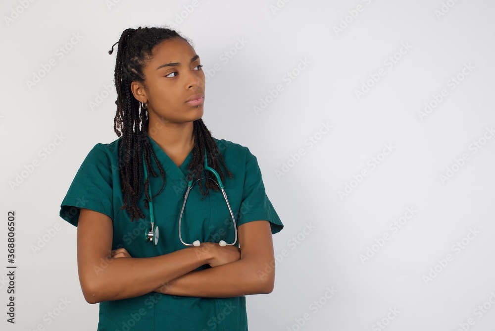 Image of upset pretty young doctor lady standing indoors with arms crossed. Looking with disappointed expression aside after listening to bad news. Confident girl. Image of upset pretty young doctor lady standing indoors with arms crossed. Looking with disappointed expression aside after listening to bad news. Confident girl.