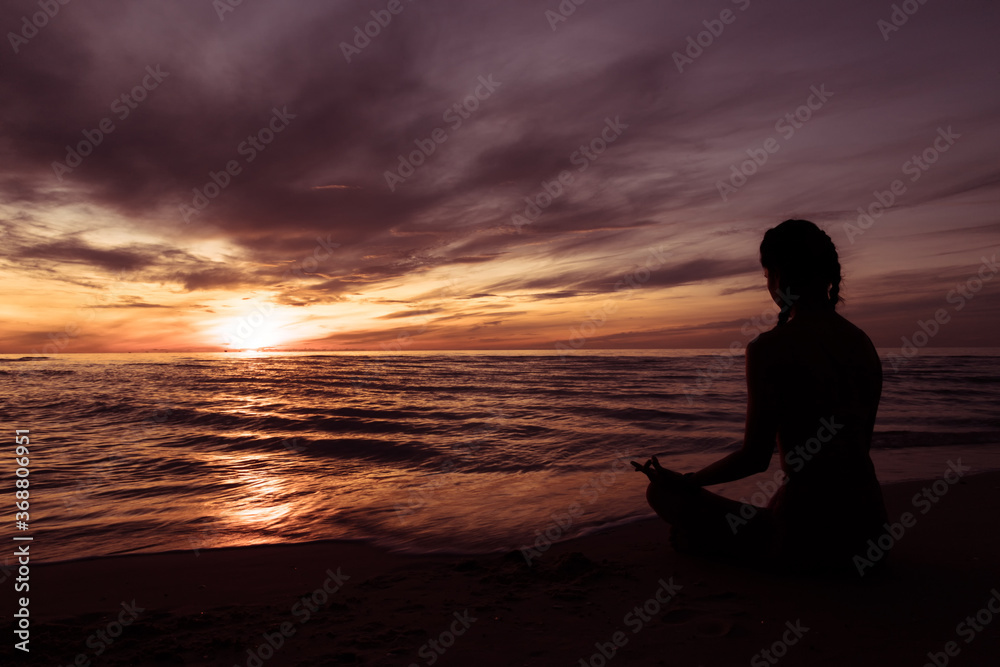 Silhouette of young woman wearing exercise clothes practicing yoga on the beach at sunset or sunrise. women do yoga. women exercising yoga in the beach.Young girl meditating in lotus posture.