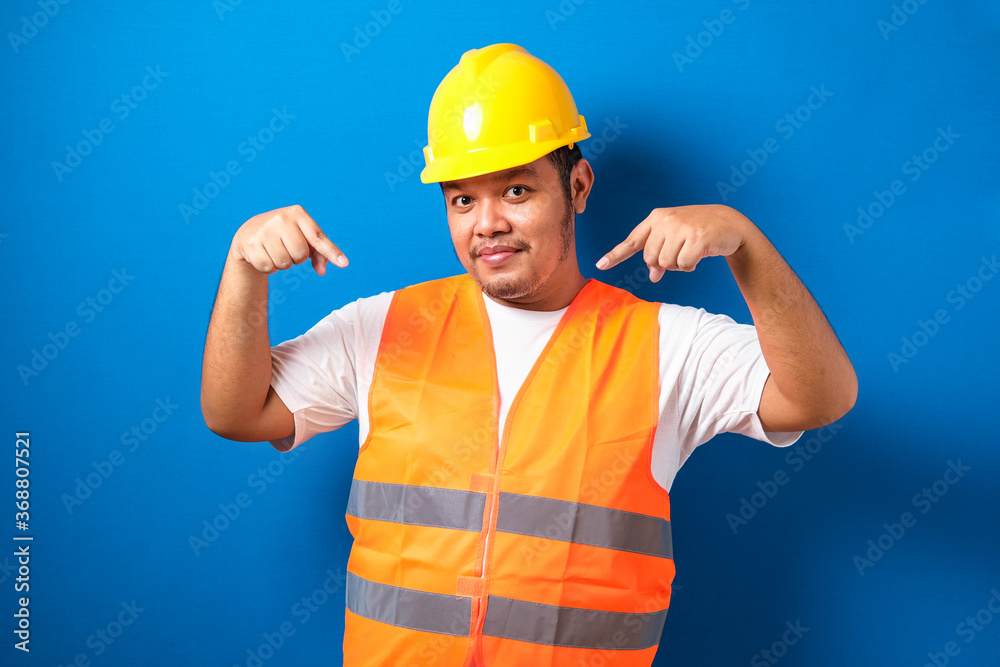 Young fat asian man over blue background wearing contractor uniform and safety helmet pointing and showing with his finger