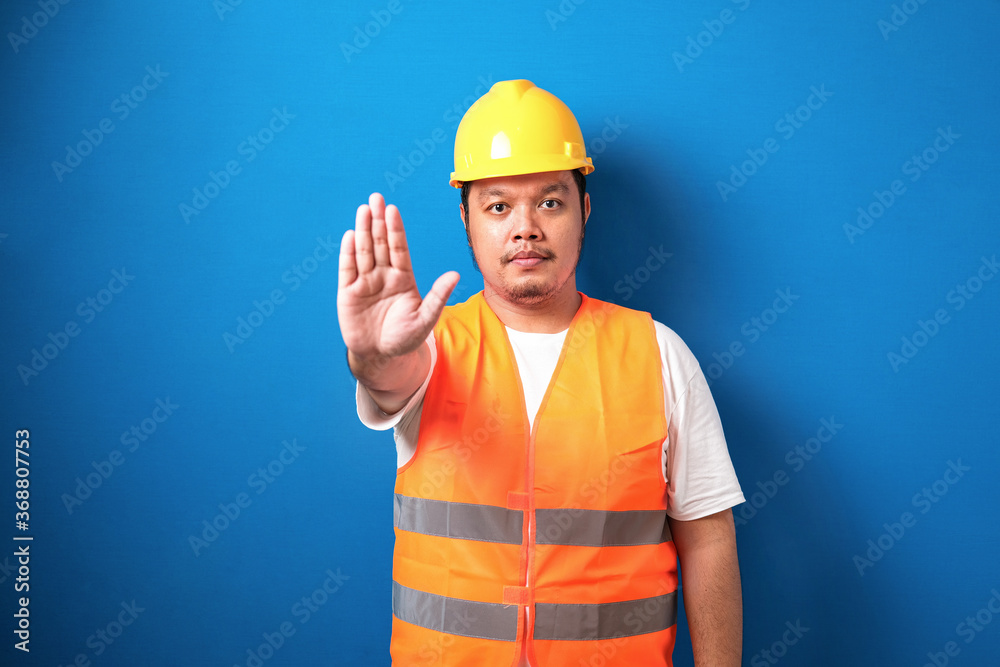 Fat asian workman wearing orange safety vest and yellow helmet making stop gesture