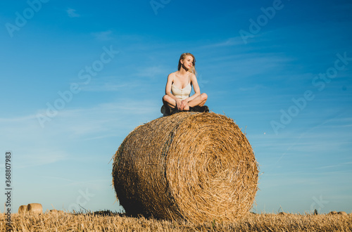 blonde girl is sitting on rolled haystack in field in sunset time