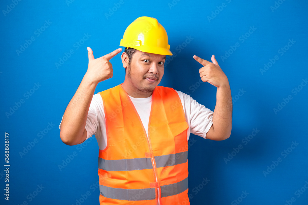 Construction worker man wearing safety vest and helmet over blue background smiling pointing to head