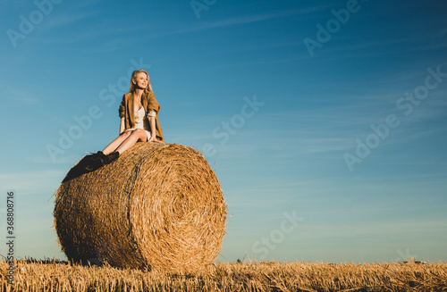 blonde girl is sitting on rolled haystack in field in sunset time