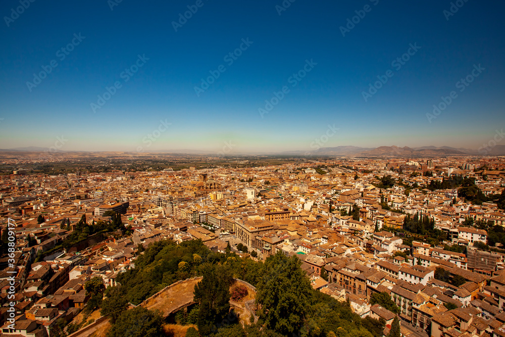 Aerial view of the historic Spanish city of Granada featuring rooftops of buildings, cathedrals, roads, trees, naked mountains in background as well as a clear blue sky.