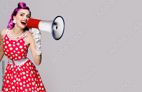 Purple head woman using megaphone, shouting something. Pin up girl in red polka dot dress. Retro vintage studio concept. Grey color background. Copy space place for some text.