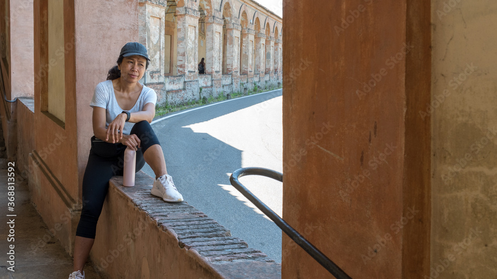 Woman in cap and sneakers holding thermos and sitting in window of portico looking at camera Woman in cap and sneakers holding thermos and sitting in window of portico looking at camera
