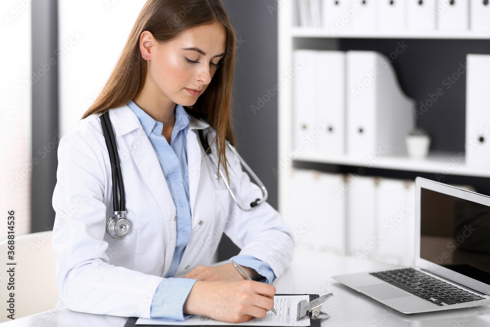 Doctor woman filling up medical form while sitting at the desk in hospital office. Physician at work. Medicine and health care concept Doctor woman filling up medical form while sitting at the desk in hospital office. Physician at work. Medicine and health care concept