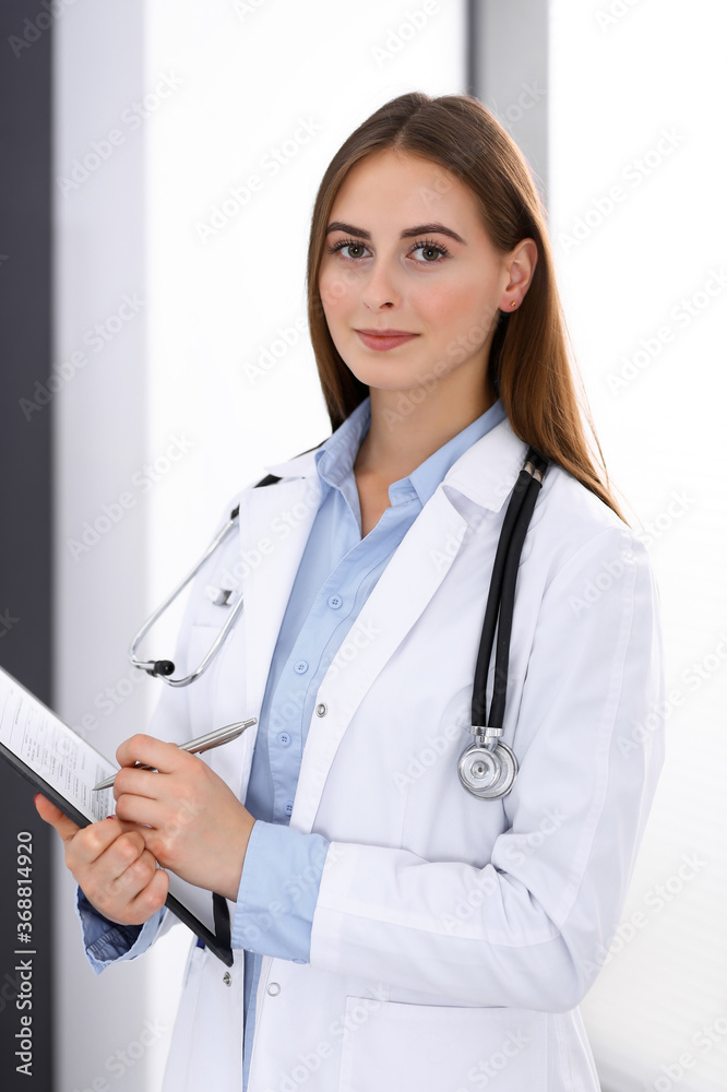 Doctor woman filling up medical form while standing near window in hospital office. Happy physician at work. Medicine and health care concept Doctor woman filling up medical form while standing near window in hospital office. Happy physician at work. Medicine and health care concept