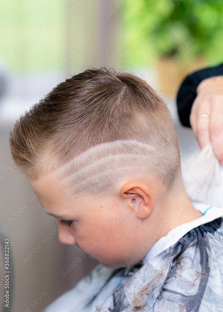 Side view caucasian american pretty school boy after haircut done stylish hairstyle wipes the hair at bright barbershop. Side view caucasian american pretty school boy after haircut done stylish hairstyle wipes the hair at bright barbershop.