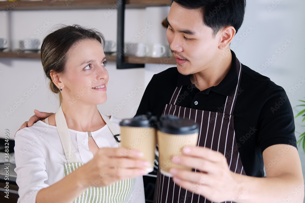 Smiling young barista couple loves Asian man and caucasian woman holding cups of hot coffee together. Start up Coffee shop and cafe business concept. Smiling young barista couple loves Asian man and caucasian woman holding cups of hot coffee together. Start up Coffee shop and cafe business concept.